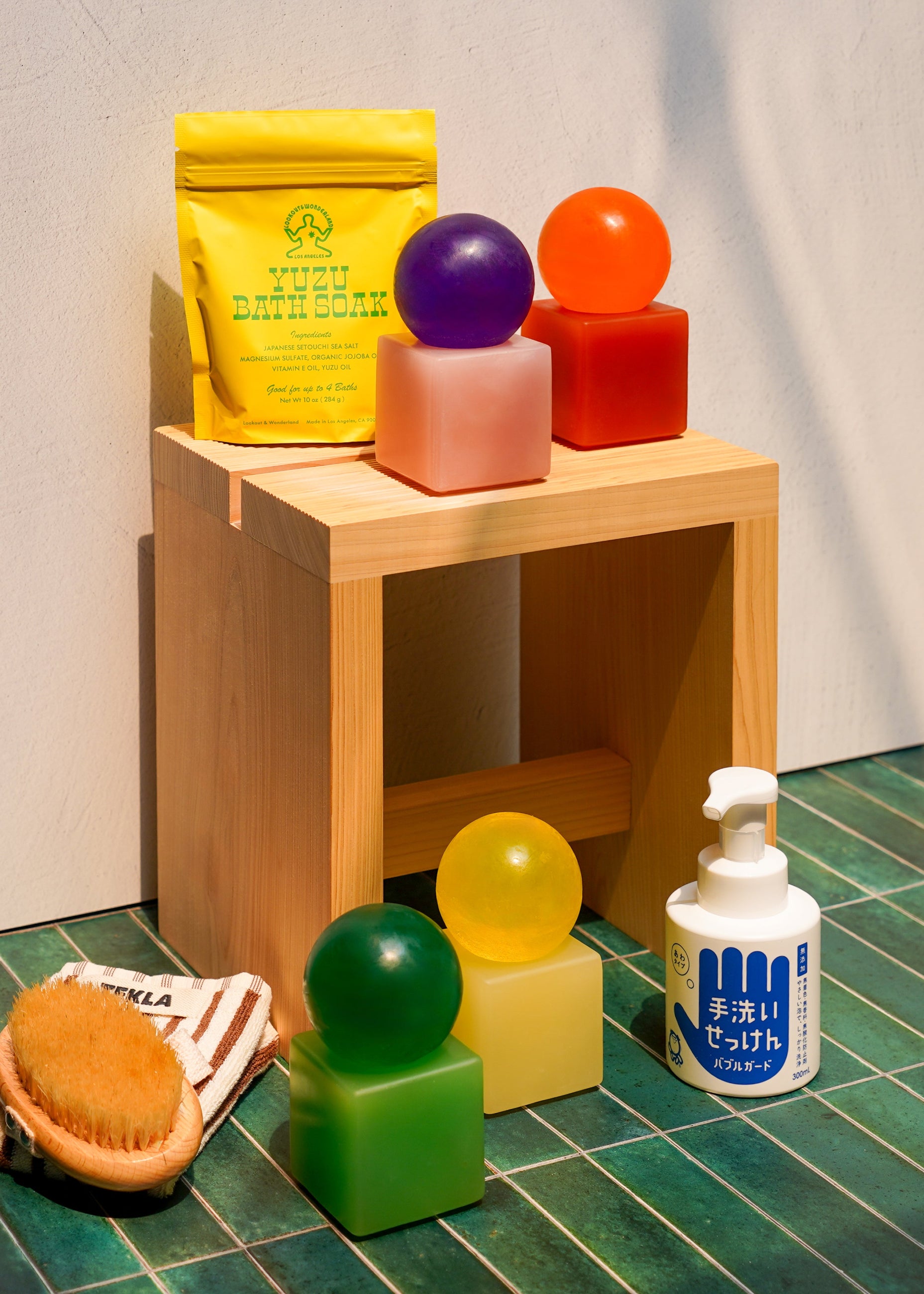 A styled product display featuring a yellow pouch of 'Yuzu Bath Soak' on a light wood Hinoki sauna stool, alongside several translucent, colorful bath soaps (purple, orange, pink, green, yellow) and a white hand soap with Japanese text on a green tiled floor.